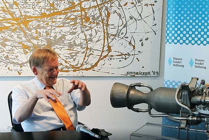 Prof. Schmucker sitzt hinter seinem Schreibtisch, linke Bildseite und gestikuliert. In der Mitte des Bildes steht das Modell eines Raketentriebwerkes vor einer HSS-Stellwand. Rechts am Bildrand sitzt Erich Kornberger in einem karierten Sacko und wartet die Antwort auf eine Frage ab. 