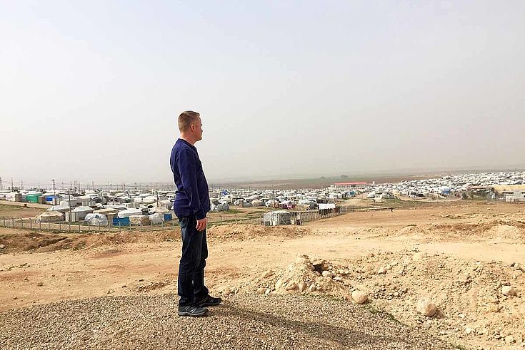 Ein Mann steht auf einem staubigen Hügel in der Wüste. Im Hintergrund die grauen, weißen Siluetten der Zeltstadt.