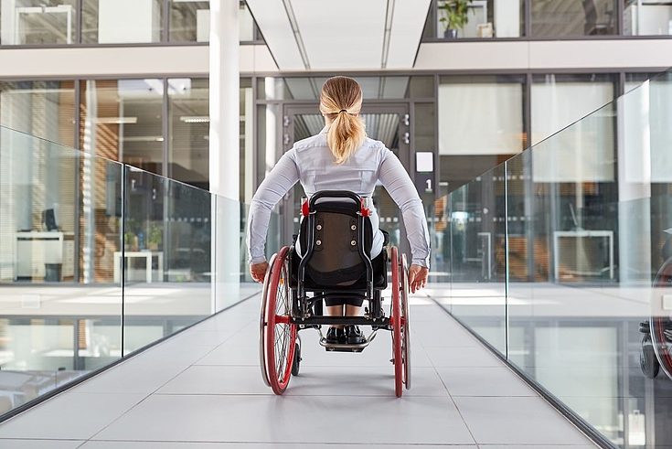 Eine Frau im Rollstuhl fährt einen modernen Gang in einem Bürogebäude entlang.