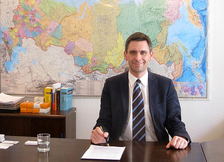 Ein freundlich blickender Mann an einem Schreibtisch mit dem Füller in der Hand. An der Wand hinter ihm eine große Weltkarte, auf der Russland hervorgehoben ist.
