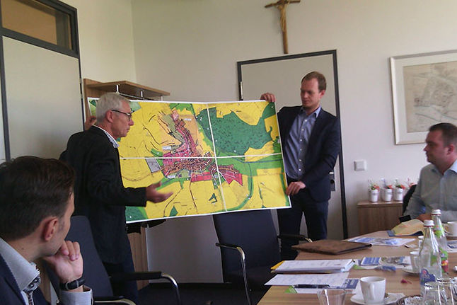 Zwei Männer halten einen Gemeindeplan so in ihren Händen. dass die Delegationsteilnehmer sie sehen können