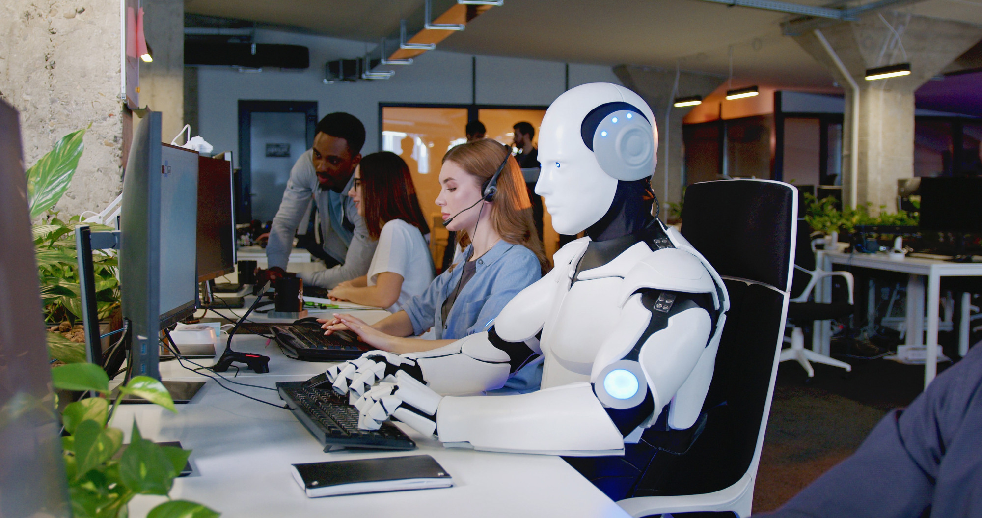 Ein Roboter arbeitet in einem Büro am Computer, daneben Personen am Computer